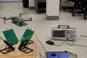 A laboratory setup on a light-colored floor showing a small gray quadcopter drone hovering just above several green rectangular panels mounted on adjustable stands fixed to a wooden base. Thin coaxial cables connect the panels to measurement equipment. To the right, a benchtop spectrum analyzer or signal analyzer with a display screen showing waveform data is connected by cables to the setup. Additional electronic components, including a small black power adapter and a blue device with a cooling fan, are placed nearby. Office chairs and desks are visible in the background, indicating an indoor lab environment.